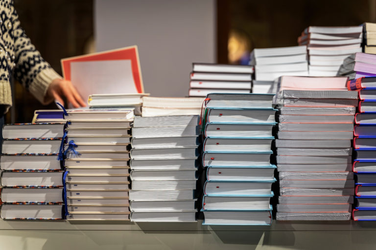Pile Of Books, Abstract Blurred Background In Book Store, In Library. Hand Of Man Leafing Through Pages Of A Book. Education, School, Study, Reading Fiction Concept.