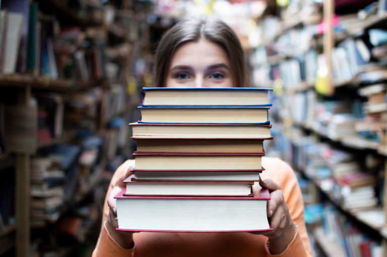Girl Student Holds A Stack Of Books In The Library, She Searches For Literature And Offers To Read, A Woman Prepares For Study, Knowledge Is Power, Bookseller On The Background Of The Bookstore, Concept